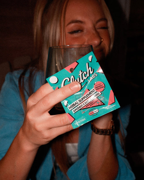 A woman in a blue top holds a glass and a colorful package of Clutch Labs Herbal Supplements. The packaging is teal with pink and white graphics, and the background is dark and dimly lit.